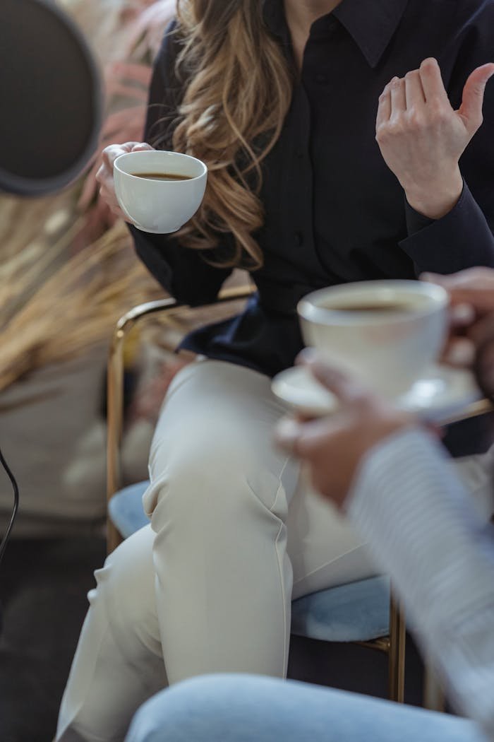 Crop anonymous female host talking to guest and having coffee while recording conversation on microphone